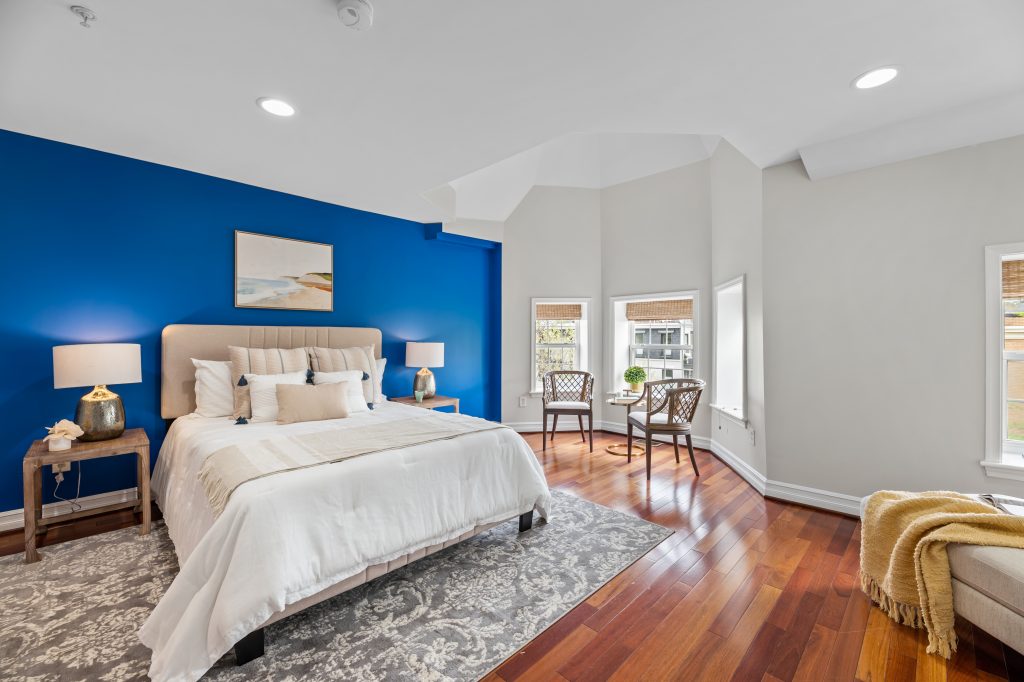 Elegant primary bedroom with bold blue accent wall, bay windows, hardwood floors, and natural light highlighting the cozy, modern staging.