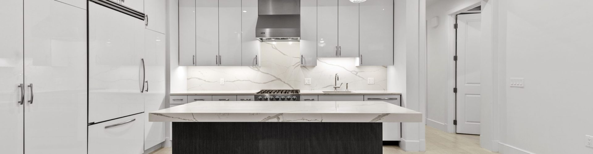 Gallery Image – Sleek White Minimalist Kitchen Design | Prismatic Home Photography Minimalist white kitchen with slab backsplash, integrated appliances, pendant lighting, and a central island with marble waterfall edge.